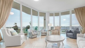 Ein geräumiges Wohnzimmer mit einem großen runden Tisch, mehreren gemusterten Sesseln und einer Couch. Das Zimmer bietet große Fenster mit Meerblick und einen Balkon.