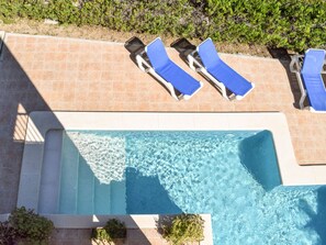 An aerial view of a swimming pool with clear water, surrounded by tiled flooring and blue lounge chairs.