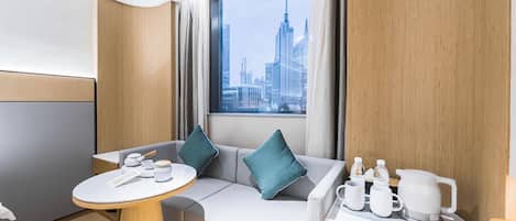 A modern hotel room with a city view, a sofa, a small table, and a kitchenette area.