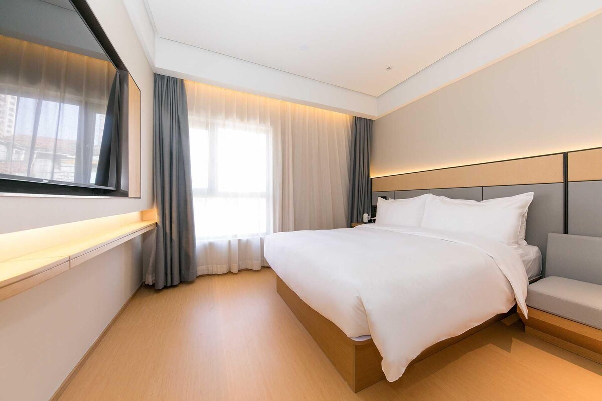 A modern hotel room with a large bed, a bedside table, a TV, and a window with curtains.