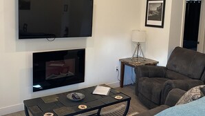 Ett vardagsrum med en platt-TV som är monterad på väggen, en öppen spis under den, ett soffbord med fjärrkontroller, en soffa och en inramad tavla på väggen.