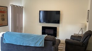 Ett vardagsrum med en mörk soffa, en fåtölj, en platt-TV som är monterad på väggen och ett litet bord med en lampa.
