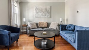 Een woonkamer met een grijze bank, twee blauwe fauteuils, een ronde salontafel en een schilderij aan de muur.