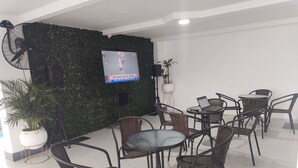 Um lobby com cadeiras de vime, uma mesa de vidro, uma TV em um suporte e uma parede verde com uma câmera.