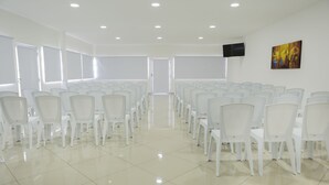Uma sala de conferências com fileiras de cadeiras brancas, uma TV de tela plana na parede e um quadro.