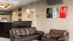 Uma área de recepção com um balcão preto, dois sofás de couro marrom, um ar condicionado fixado na parede e uma pintura de árvores.