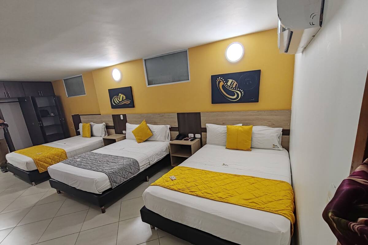 Ein Hotelzimmer mit drei Betten, jedes mit einer gelben Decke, weißer Bettwäsche und einem Holz-Kopfteil. Es gibt einen Kleiderschrank, einen Schreibtisch mit Stuhl und einen kleinen Tisch mit einer Lampe.