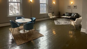 A spacious living room with a round table, blue chairs, a sofa, and a coffee table. There are large windows with floor-to-ceiling views and wall-mounted lights.