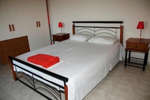 Cama bem arrumada com lençóis brancos e toalha vermelha, mesa de cabeceira de madeira com abajur vermelho e guarda-roupa com duas portas.