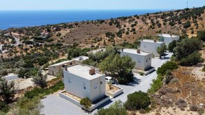 A coastal property with three white buildings, each with a flat roof and a small garden area in front. The buildings are surrounded by dry vegetation and a hillside in the background. - Paterakis Big Studio in Triopetra (Agios Vasileios)