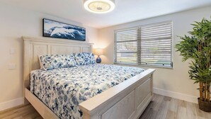 Ein Schlafzimmer mit einem Bett, einer Deckenleuchte, einem Fenster mit Jalousien und einer Zimmerpflanze.