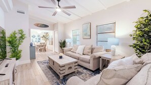 Ein geräumiges Wohnzimmer mit Deckenventilator, einer beigefarbenen Sofagarnitur, einem Couchtisch und einem Essbereich im Hintergrund.