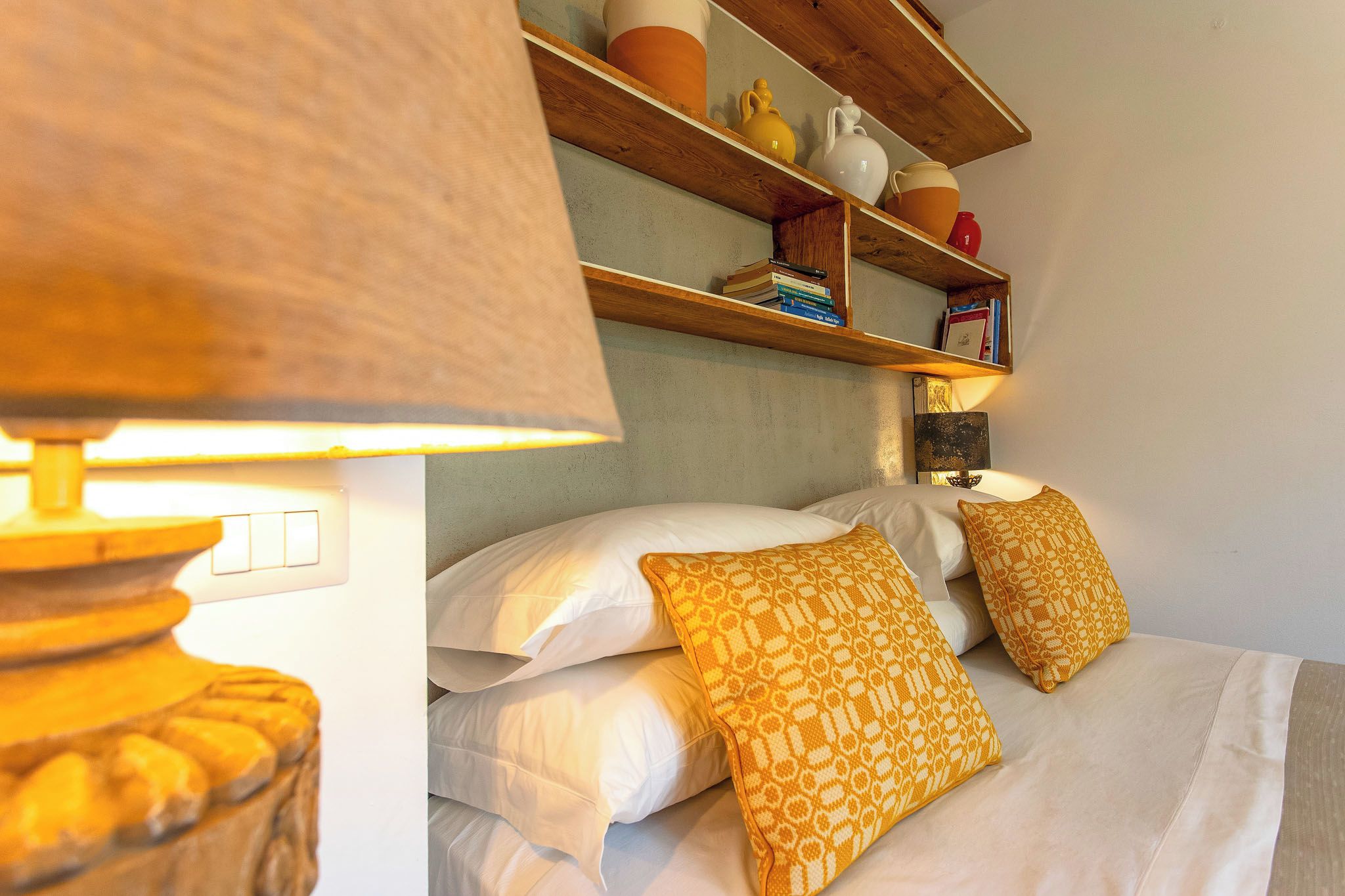 Un lit avec une literie blanche et des oreillers à motifs, une lampe et une étagère avec des livres et des objets décoratifs.