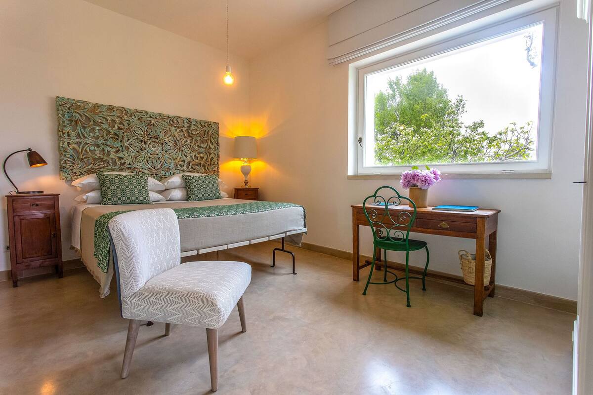 A bedroom with a bed, bedside lamps, a desk, a chair, and a window with a view of trees.