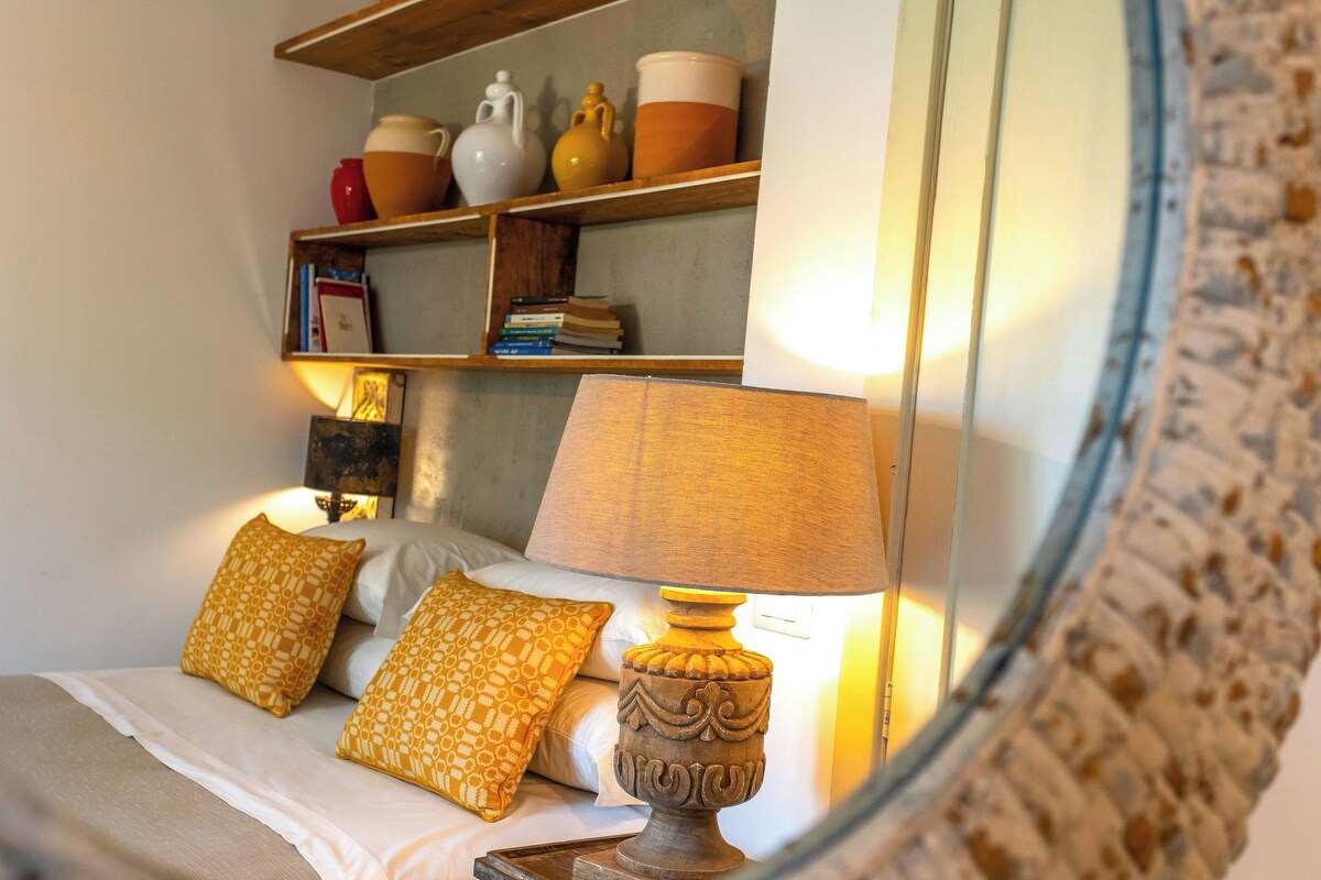 A bedroom with a bed, two patterned pillows, a bedside lamp, a shelf with books and decorative items, and a mirror reflecting part of the room.