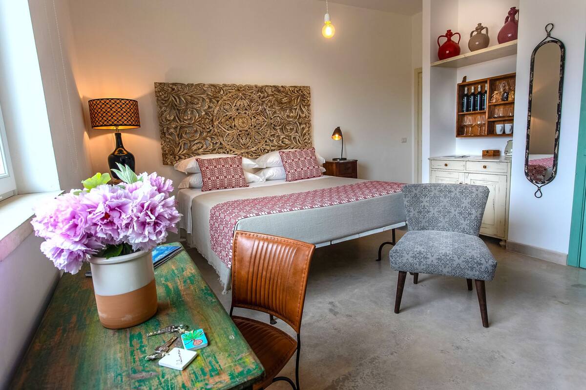 A bedroom with a bed, bedside lamps, a chair, a table with a vase of flowers, and a dresser with decorative items.