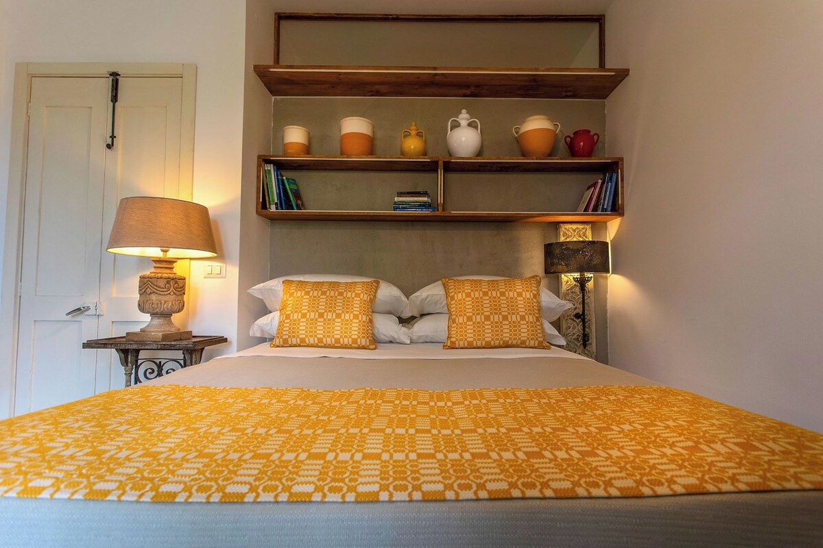 A neatly made bed with a patterned bedspread, a bedside lamp, and a shelf with books and decorative items.
