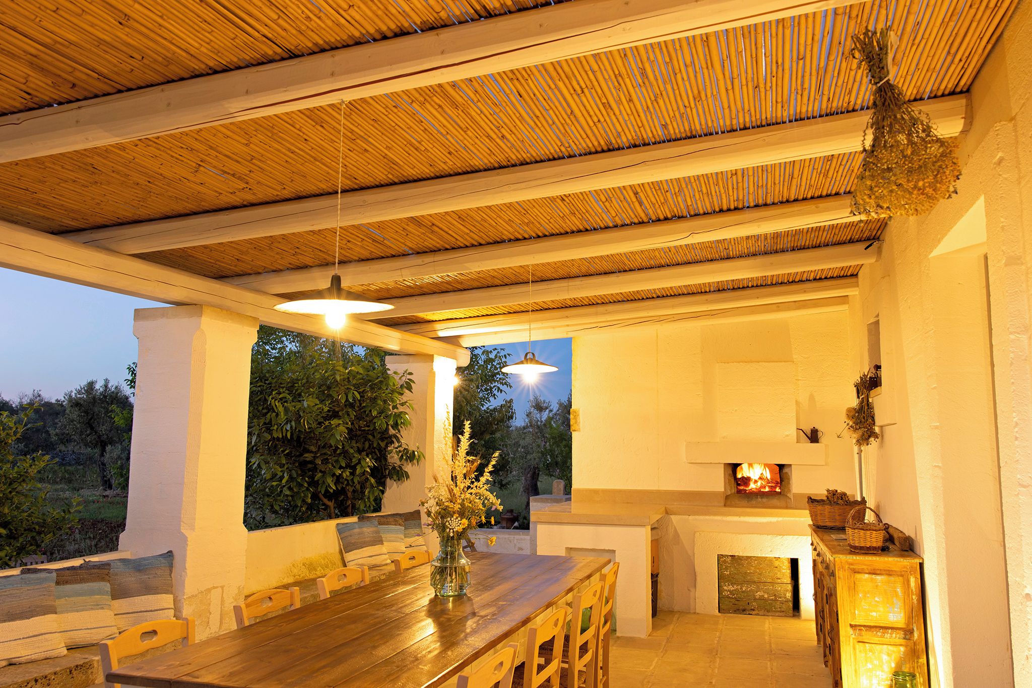 Un espace extérieur aménagé pour les repas, avec une table et des chaises en bois, un luminaire suspendu et une cheminée.