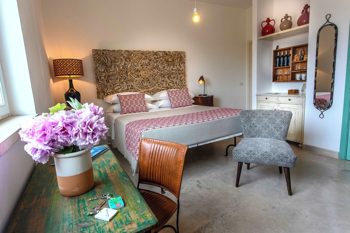 A bedroom with a bed, bedside lamps, a chair, a table with a vase of flowers, and a dresser with decorative items.