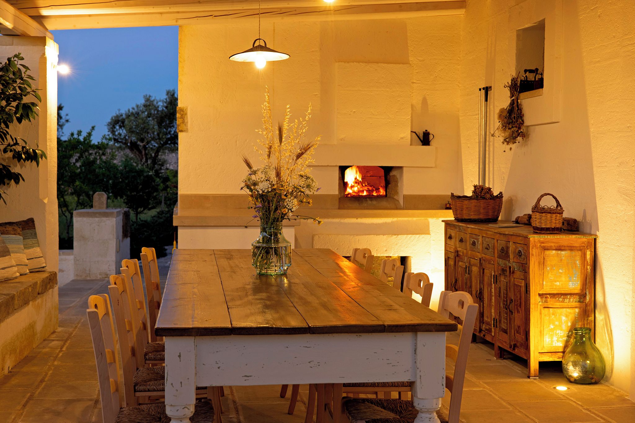 Un espace extérieur aménagé pour les repas, avec une table et des chaises en bois, un vase rempli de fleurs et une cheminée en arrière-plan.