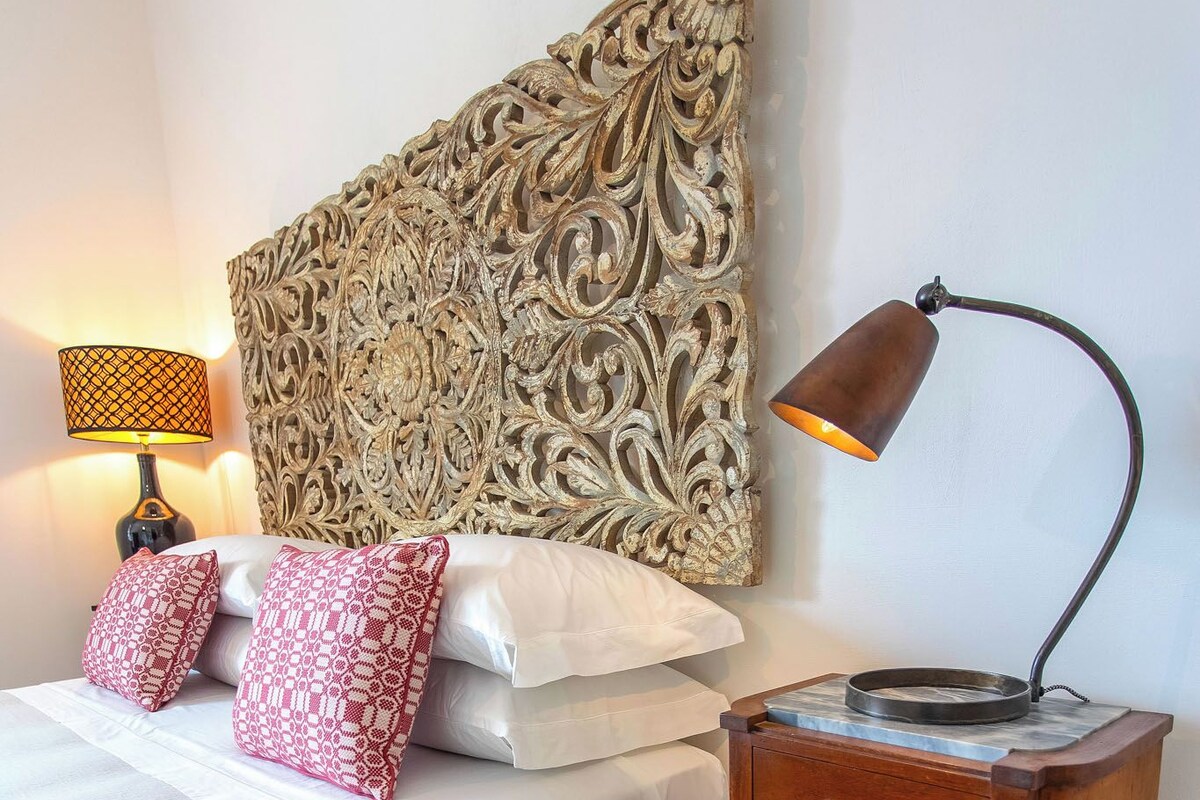 A neatly made bed with white linens and patterned pillows, a decorative wall headboard, a bedside lamp, and a small wooden nightstand with a round table.