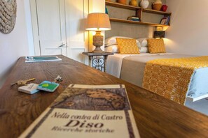 A hotel room with a wooden table, a lamp, a bed with patterned bedding, and a shelf with books and decorative items. - Masseria Ferarelli Trepizzi (Diso)