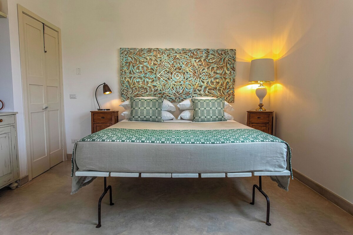 A neatly arranged bedroom with a bed, two bedside tables, a decorative wall hanging, and a wall-mounted lamp.