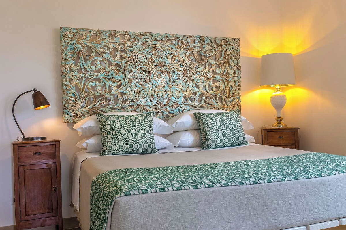 Un lit bien fait, agrémenté d’oreillers à motifs, d’une tête de lit décorative, d’une lampe de chevet et d’une petite table de nuit en bois.