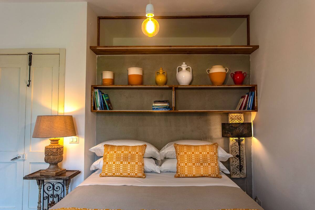 A bedroom with a bed, two bedside lamps, a shelf with decorative items, and a hanging light fixture.
