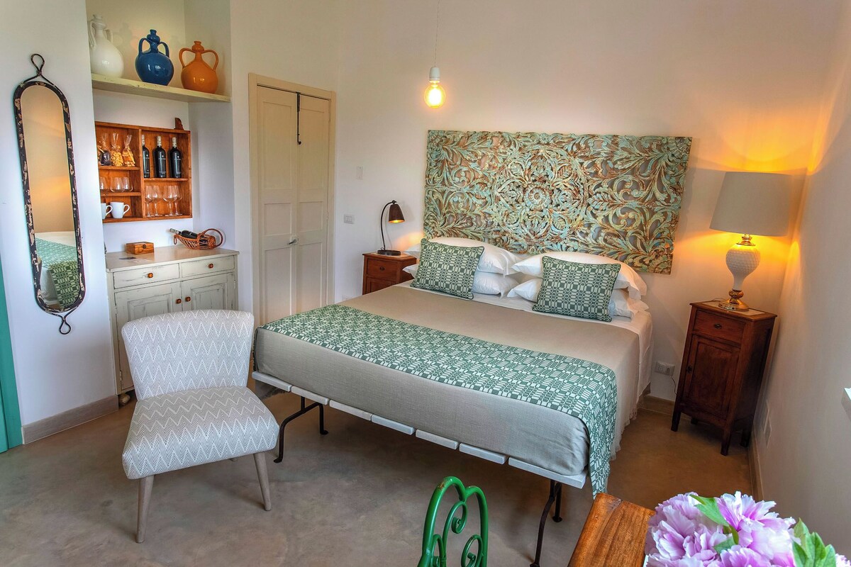 A bedroom with a bed, a chair, a nightstand, a lamp, a mirror, shelves with bottles and glasses, and a door.