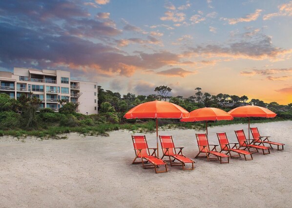 Une plage avec des parasols rouges et des chaises longues, un bâtiment en arrière-plan et un ciel au coucher du soleil.