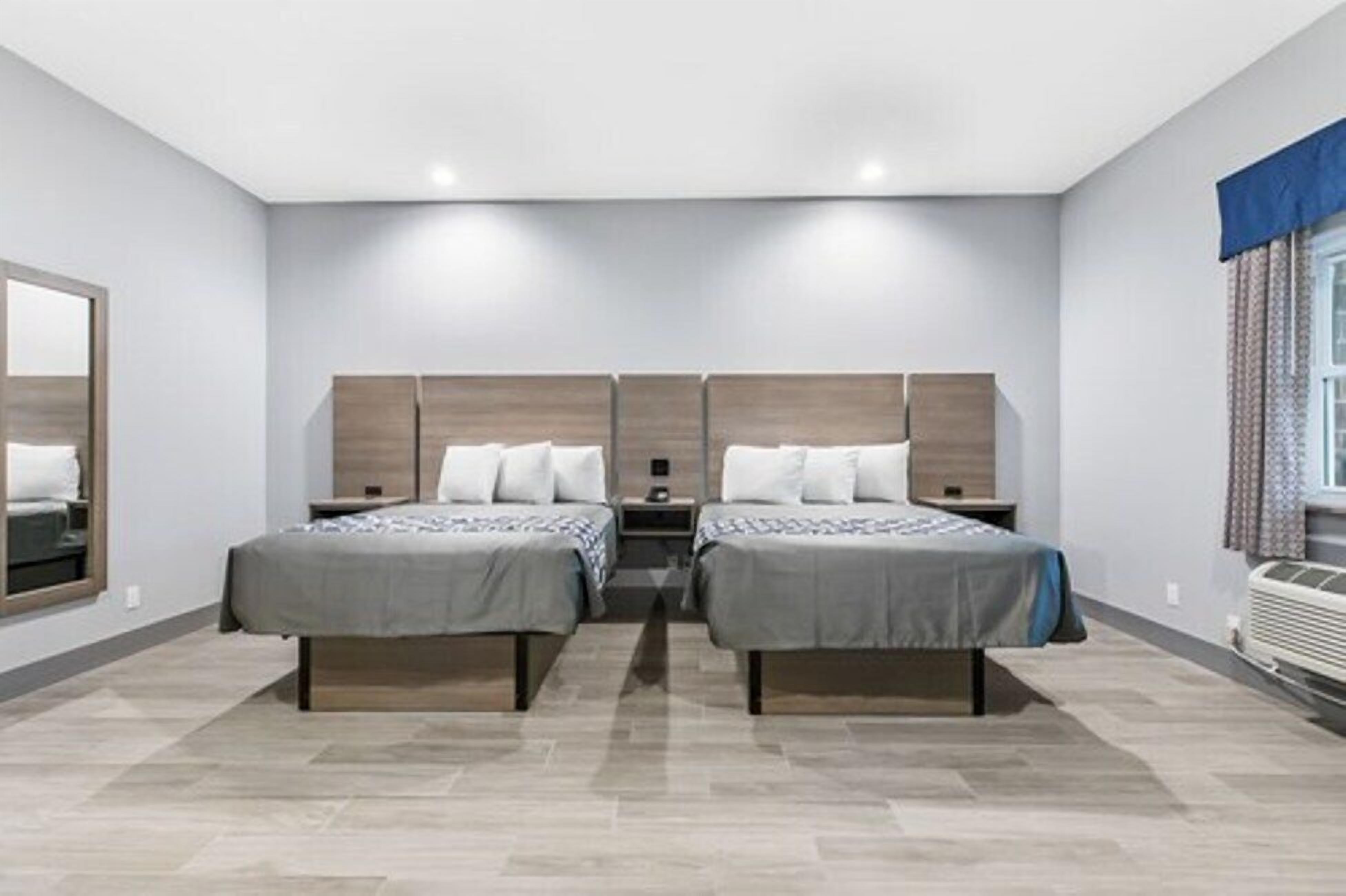 A hotel room with two beds, a large mirror, a window with curtains, and a built-in air conditioning unit.