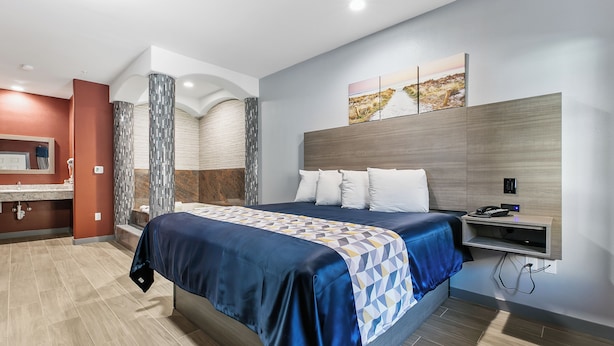 Ein modernes Hotelzimmer mit einem großen Bett, einem Nachttisch, einem integrierten Fernseher sowie einem Badezimmer mit Dusche und Toilette.