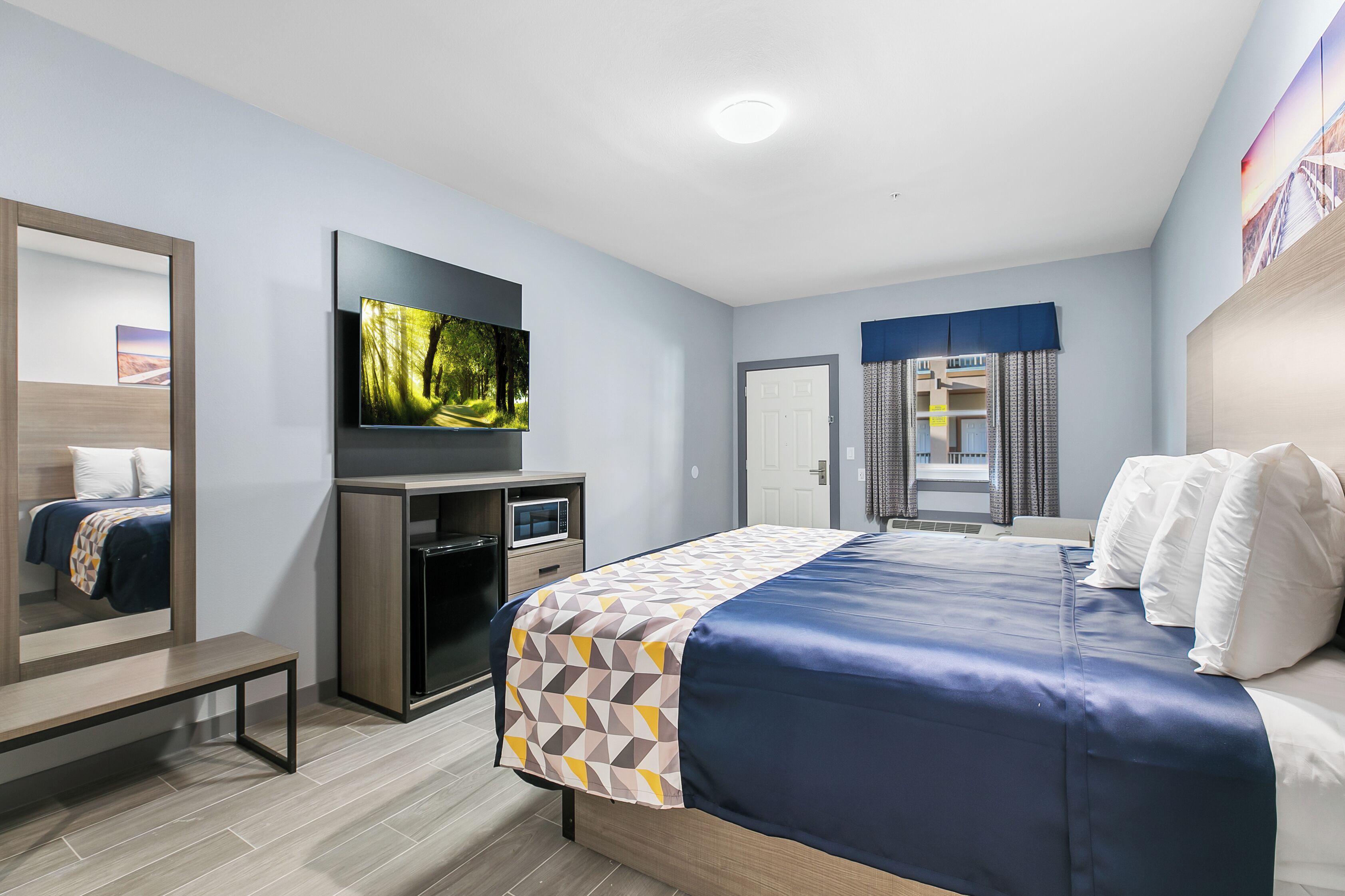 A modern hotel room with a large bed, a flat-screen TV mounted on the wall, a desk with a computer, and a mirror.
