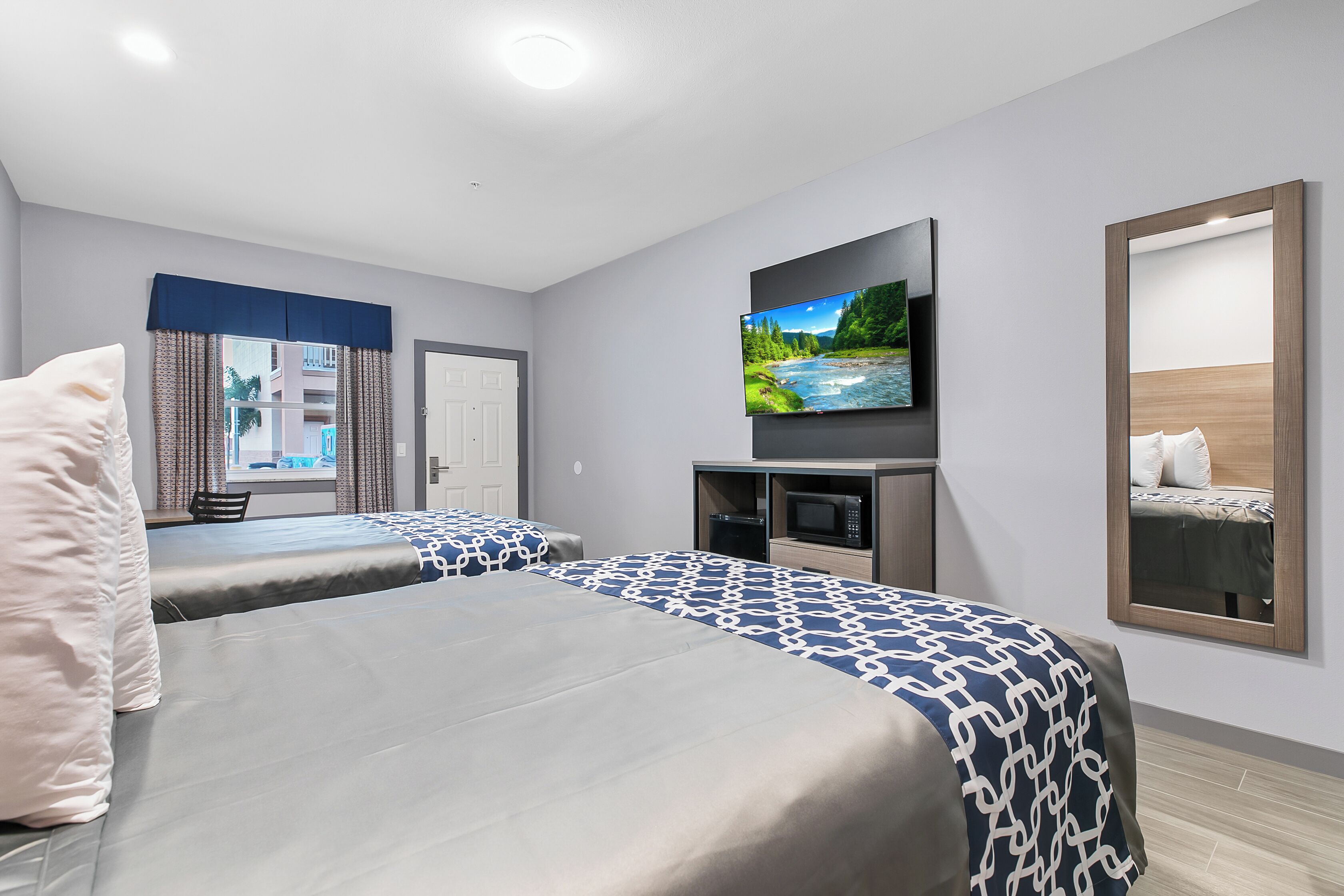 A hotel room with two beds, a TV, a mirror, and a view of a building outside.