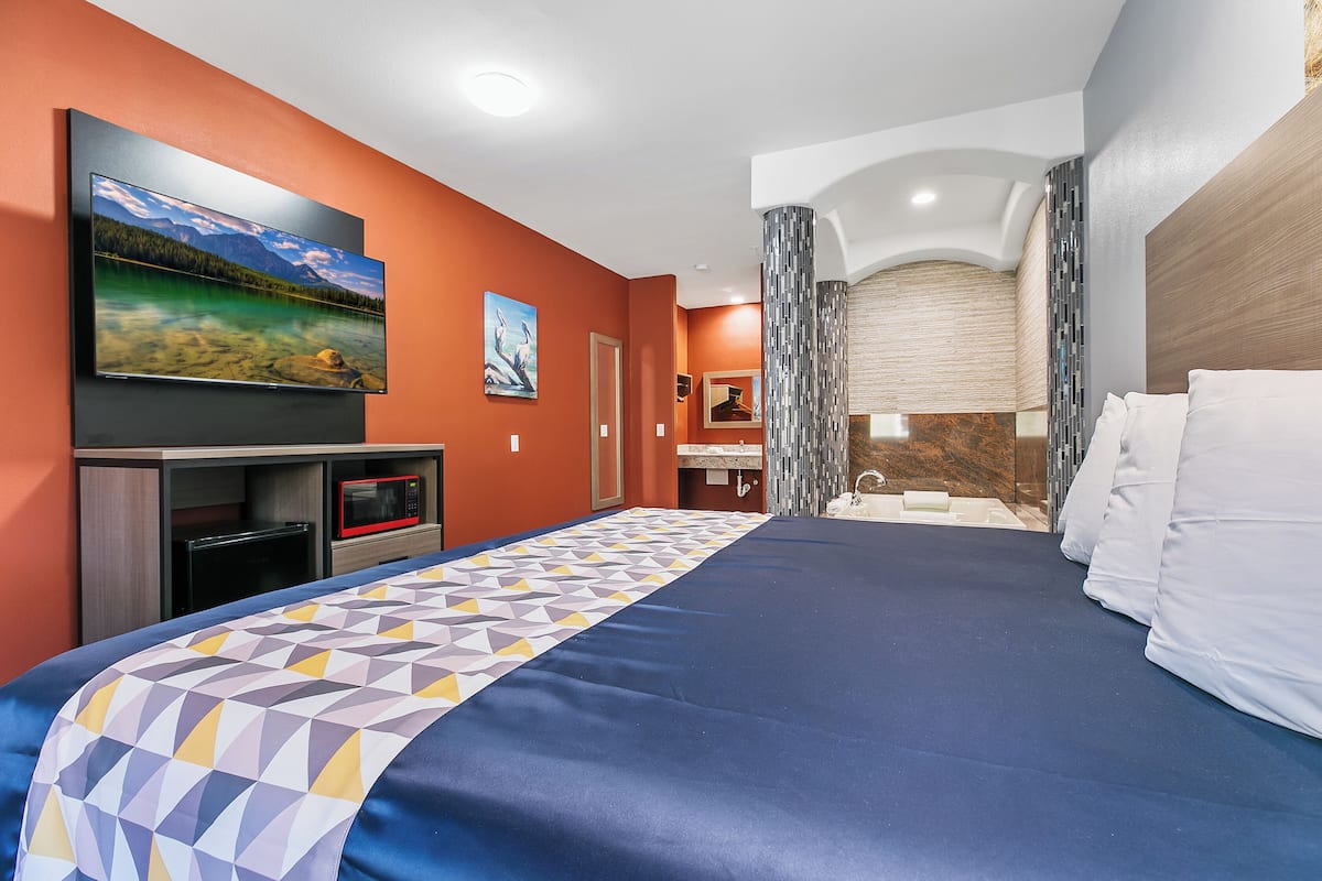 Habitación de hotel con una cama grande, un televisor de pantalla plana montado en la pared, un microondas y una pequeña cocina.