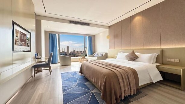 Una habitación de hotel moderna con una cama grande, un área de descanso con vistas y un paisaje urbano visible a través de la ventana.