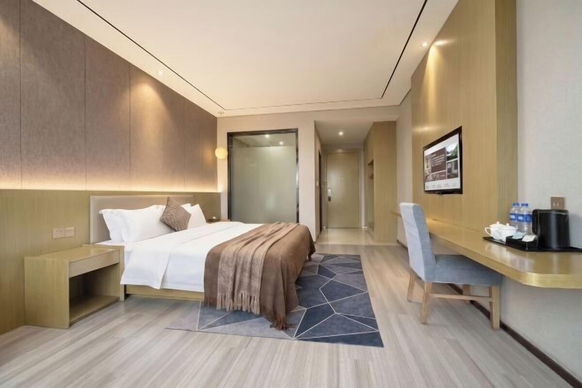 A modern hotel room with a bed, bedside table, desk, chair, and a view of the hallway.