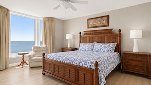 A bedroom with a large bed, a chair, a nightstand, a bedside table, a ceiling fan, and a window with curtains.