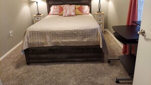 A neatly made bed with a wooden headboard, two bedside tables with lamps, a small desk with a chair, and a red curtain. - Chateau Rose (Branson)