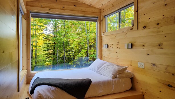 Un dormitorio tipo cabaña de madera con una ventana grande, una cama y vistas a un bosque.