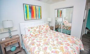 A bedroom with a bed, a nightstand, a lamp, a mirror, and a colorful painting on the wall. - Dog Friendly Beach House, Steps to Beach, Ocean View Sleeps 10! (Freeport)