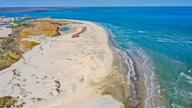 Vista aérea de una zona costera con una playa de arena, un pequeño asentamiento con edificios y un océano azul cristalino.