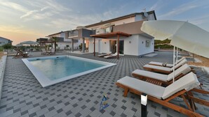 A swimming pool with lounge chairs and umbrellas, a covered patio area, and a modern house with a balcony. - Modern 4 BD villa with heated pool for up to 8 guests (Sukošan)