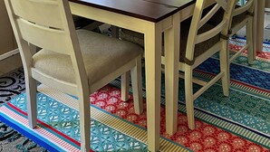 Una mesa de comedor con seis sillas, un repuesto a cuadros sobre las ventanas y una alfombra colorida en el suelo.