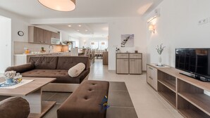 A modern living room with a sofa, ottoman, and a TV on a wooden stand. There is a kitchen in the background with wooden cabinets and a dining area. - Exquisite 4 BD villa for 8 with heated pool near Zadar (Sukošan)