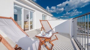 Un balcone con una sedia di legno e un tavolino, una porta scorrevole in vetro che dà su una camera da letto, e un cielo azzurro terso.