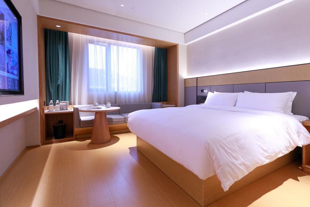 A modern hotel room with a large bed, a small table with a chair, a sofa, and a TV mounted on the wall.
