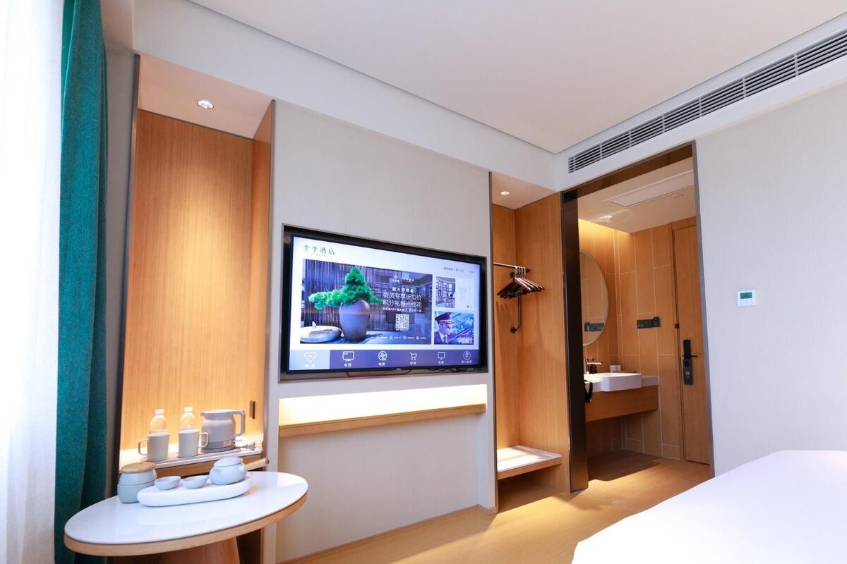 A modern hotel room with a bed, a TV mounted on the wall, a small table with cups and a tray, and a wooden wardrobe.