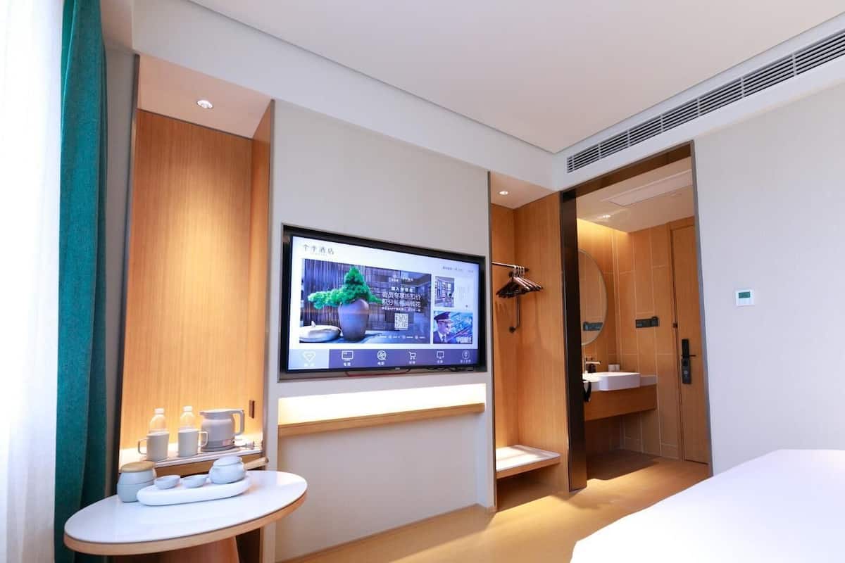 A modern hotel room with a bed, a TV mounted on the wall, a small table with cups and a tray, and a wooden wardrobe.